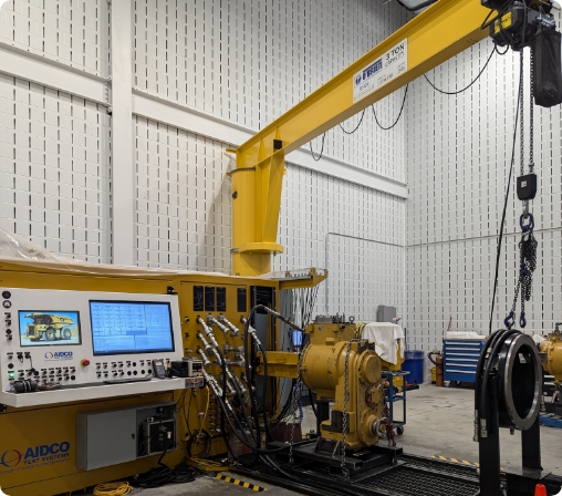 Industrial testing station featuring a yellow crane, hydraulic machinery, and a control panel with multiple monitors and cables in a well-lit workshop